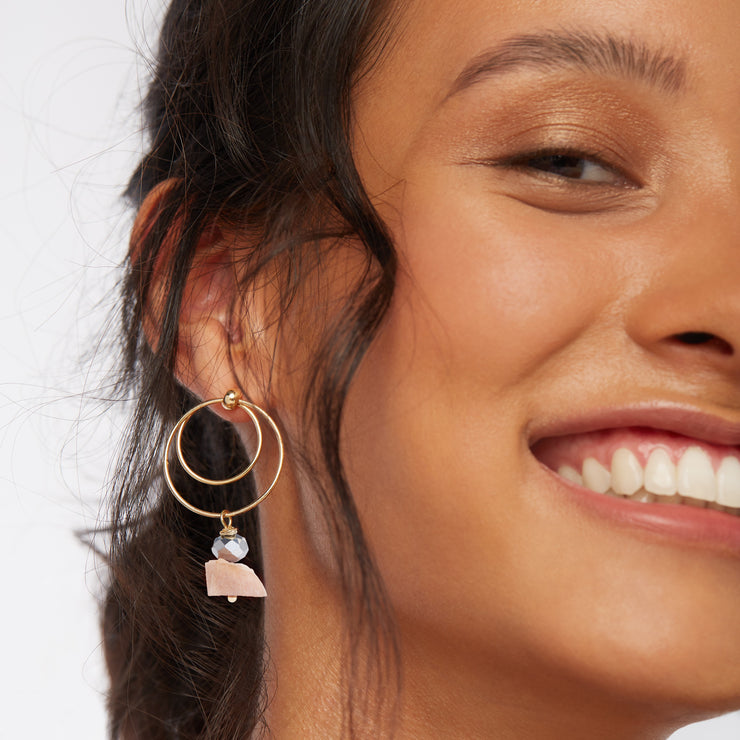 Smiling brunette wearing a pair of earrings, gold tone front facing double hoops, small and big circle, with a silver tone plated crystal cut glass and raw sunstone dangle.