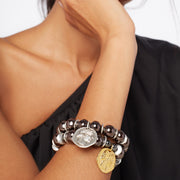 Woman's wrist. She wears a black one shoulder top and a two bracelet stack with signature Greek ceramic beads and coins-mixed metal and glossy 